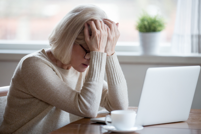 Een vrouw met een vermoeide uitdrukking, leunend op haar handen, achter een laptop. Een kop koffie staat op tafel.