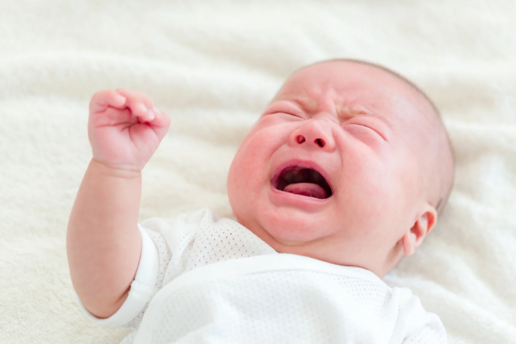 Huilende pasgeboren baby in een wit rompertje op een zachte ondergrond.