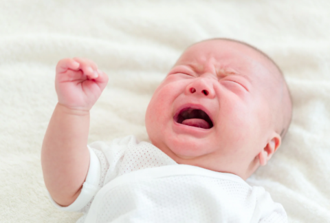 Huilende pasgeboren baby in een wit rompertje op een zachte ondergrond.