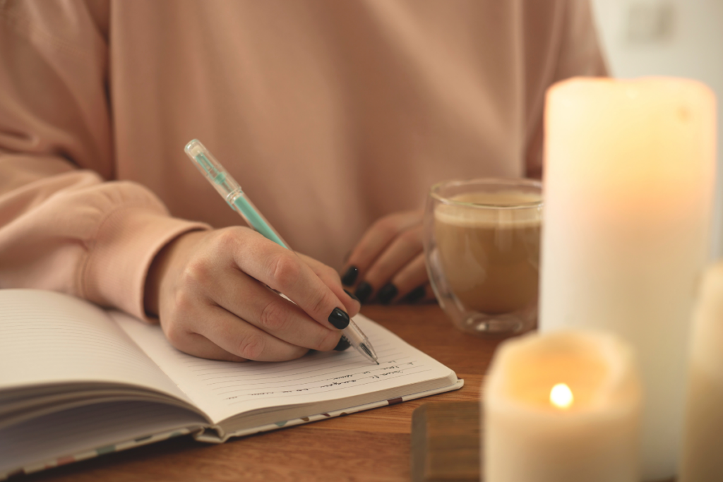 en vrouw schrijft in een journal met een turquoise pen terwijl ze geniet van een kop koffie. Kaarsen branden op de voorgrond, wat een rustgevende sfeer creëert.