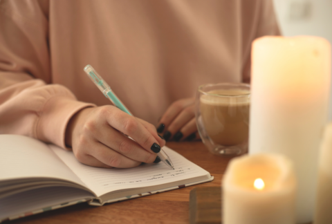 en vrouw schrijft in een journal met een turquoise pen terwijl ze geniet van een kop koffie. Kaarsen branden op de voorgrond, wat een rustgevende sfeer creëert.