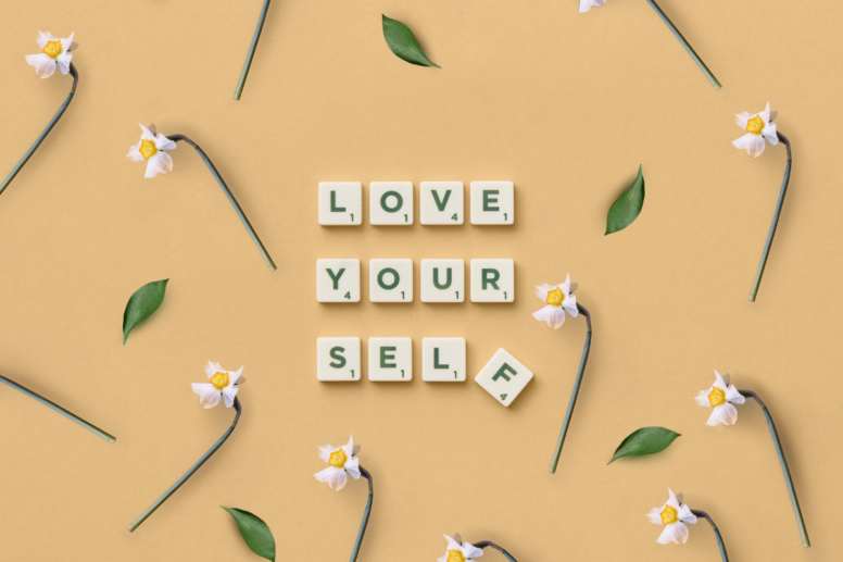 Scrabble letters die de tekst: 'love yourself' vormen op een lichter achtergrond met bloemen